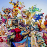 Elaborate towering falla sculpture from Fallas Valencia 2026, showcasing a celestial-themed composition with goddesses, fairies, warriors, colorful serpents, astrological symbols, and ornate costumes in vivid blues, pinks, golds, and reds, displayed in a sunny Valencia square.