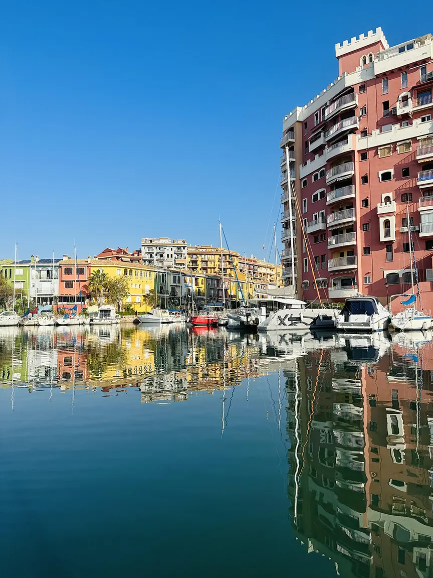 Tranquil lake reflects sky, boats draped blue, red-white blend. Cityscape with tall buildings, balconies. Harbor adds depth to serene scene. | Port Saplaya