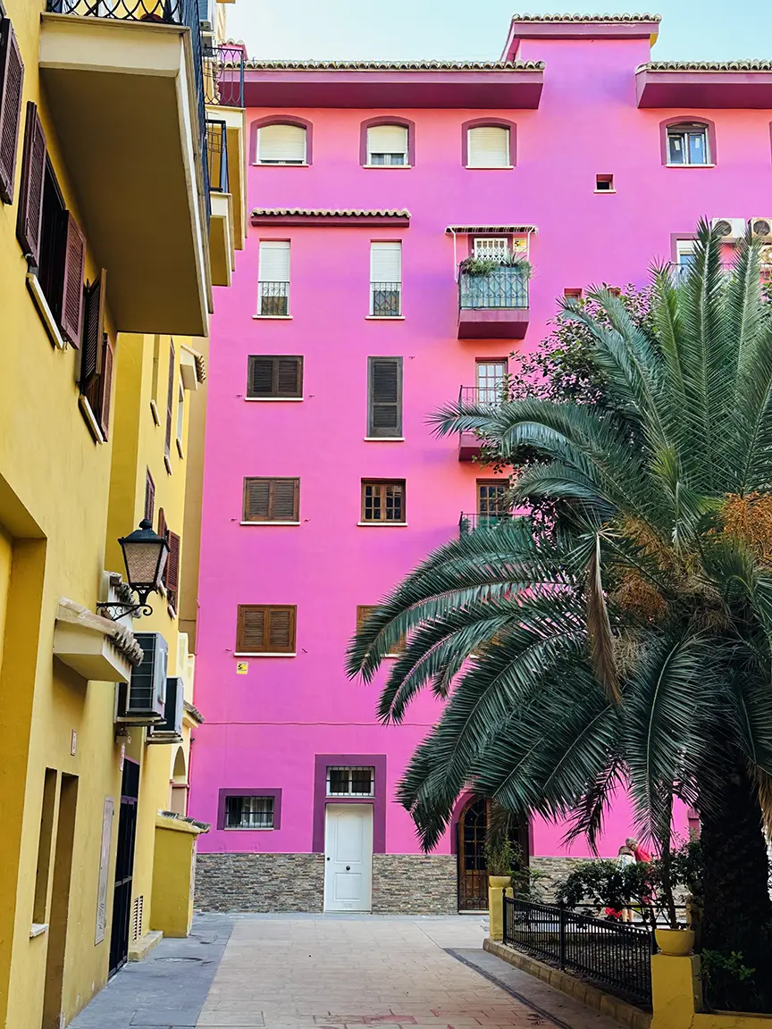 Pink building stands out against black backdrop. Windows adorned with white blinds. Palm trees add tropical vibe, softening urban lines. Black railing offers contrast. Vacation-like hotel feel. Urban meets nature in captivating spectacle. | Port Saplaya